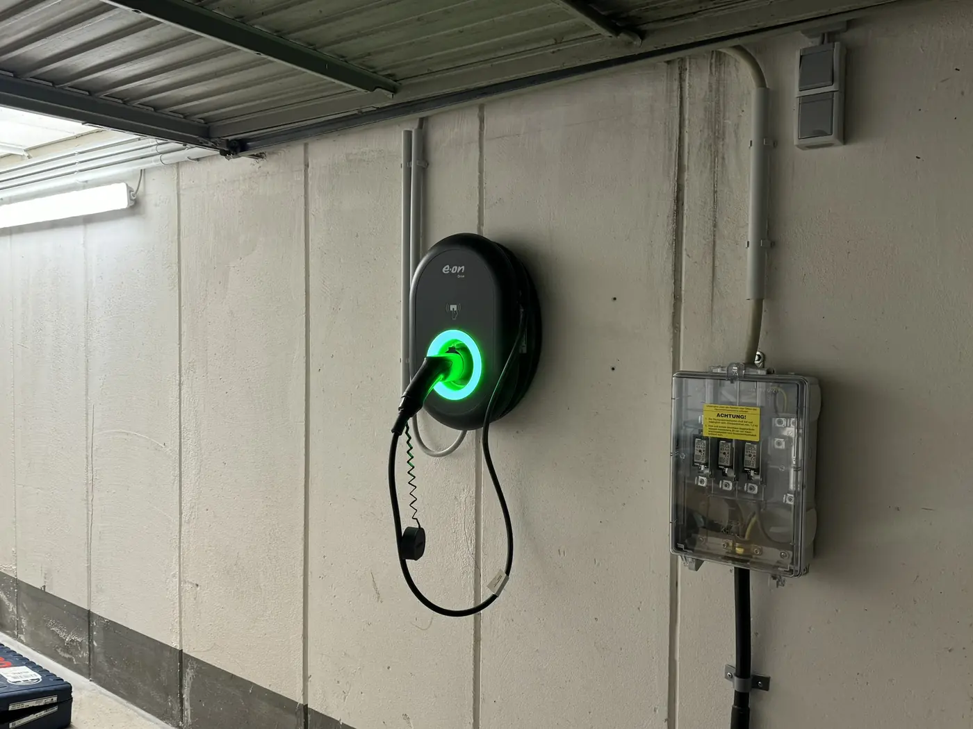 Wallbox mit grün leuchtendem Ladering an der Wand einer Garage, daneben die Absicherung